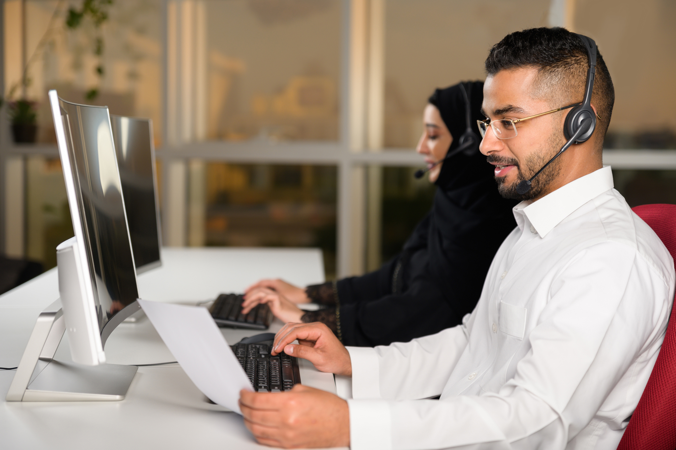 portrait of middle east male call center or help desk or telephone support agent working at call center workplace wearing headset with blurred background of other agents working; Shutterstock ID 2215768287; purchase_order: -; job: -; client: -; other: -